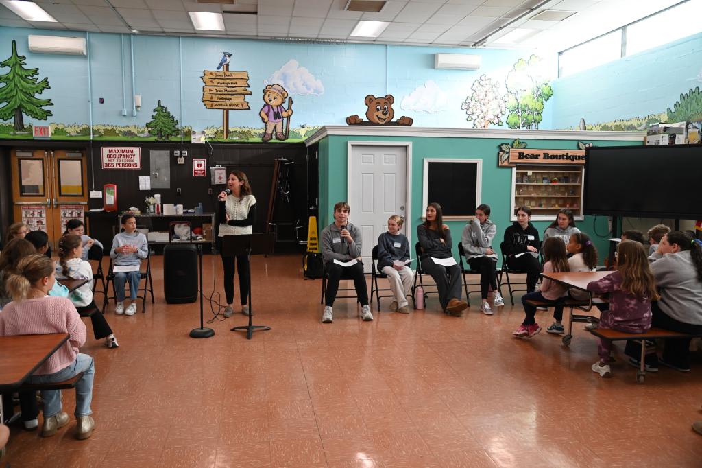 A woman speaks into a microphone while standing before a group of young people seated in chairs and at tables in a brightly painted room.